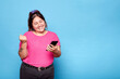 © Lux Images - Young curvy latina woman celebrating good news with a raised fist while holding her mobile phone. Indoor studio shot, isolated against a blue background.