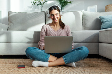 Wall Mural - Pretty young woman listening music with headphone while working with laptop sitting on the floor at home.