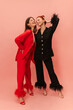 © Look! - Full-length two stylish young caucasian girls are smiling with eyes closed, posing on pink background. Models brunette and redhead are wearing fancy suits in red and black. Good mood, trends concept