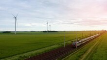 Aerial View Of Local Train Traveling Through Green Fields Under Sunset Sky And Giant Wind Turbines. Train, Railway In The Meadow, Windmills In The Distance, Sunset Sky, Drone 4K. High-speed Train. 