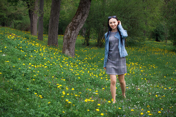 A positive, beautiful girl in a gray dress and a blue jacket, with glasses on her hair, walks through a meadow with grass and small white and yellow flowers during the day.