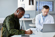 © Seventyfour - Black military officer sitting at desk signing medical service agreement while Caucasian doctor texting something on laptop during appointment