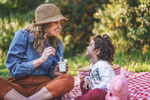 A Young Single Mother Spoon Feeds Yogurt To Her Little Daughter Sitting On The Grass During A Picnic Outdoors. Concept Of Family, Outdoor Life And Separated Parents.