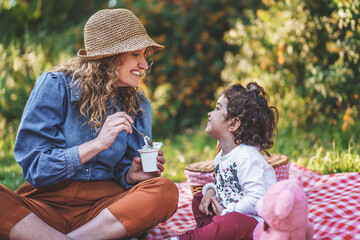 a young single mother spoon feeds yogurt to her little daughter sitting on the grass during a picnic