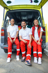 Canvas Print - Multi-ethnic group of paramedics standing at the side of an ambulance with open doors. Their coworker carrying a medical trauma bag. They are smiling at the camera.