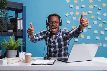 Wall Mural - overjoyed smiling call center operator showing thumbs up gesture with hands while looking at camera.