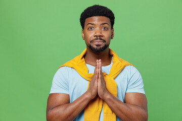 Wall Mural - Young man of African American ethnicity 20s wear blue t-shirt hold hands folded in prayer gesture, begging about something isolated on plain green background studio portrait. People lifestyle concept.