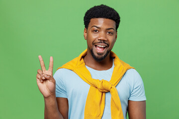 Wall Mural - Young smiling happy joyful fun cool satisfied man of African American ethnicity 20s wear blue t-shirt showing victory sign isolated on plain green background studio portrait. People lifestyle concept.