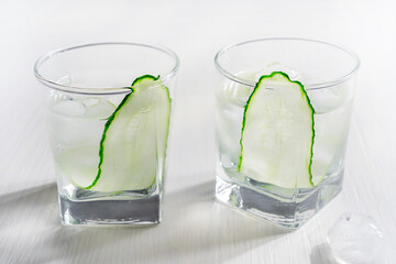 Two glasses of cold refreshing homemade cucumber water garnished or decorated with vegetable slice served with ice cubes on white wooden background as healthy summer drink full of antioxidants