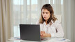 © lenblr - Disabled woman freelancer with infantile cerebral paralysis talks on videocall via laptop with manager showing project paper against window at home, sunlight.