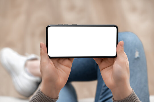 Wall Mural -  - Top view mockup image of woman holding mobile phone horizontally with blank white screen. woman using cellphone horizontal with while sitting cross-legged on sofa at home. Mockup smartphone