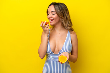 Wall Mural - Young caucasian woman isolated on yellow background in swimsuit and holding an orange
