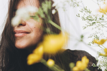 Beautiful sensual woman portrait in sunlight among summer wildflowers in room. Young brunette woman with flowers posing in sunlight. Skin care and self care