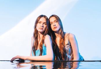 Beauty portrait with water of young beautiful girls fashion models twins sisters with healthy dark long hair on blue background