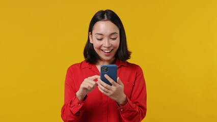 Wall Mural - Dreamful pensive minded young woman of Asian ethnicity 20s years old wears red shirt hold using mobile cell phone typing browsing chatting send sms isolated on plain yellow background studio portrait