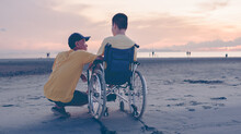 Behind Of Father Or Volunteer Or Caregiver Talking To Young Man With Disability On The Sea Beach At Sunset, Travel And Vacation In Summer, Positive Photos Give Life Power, Mental Health Concept.