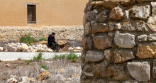 An Old Man In A Beret Sits In Front Of A Stone Wall In A Small, Uninhabited Spanish Village  