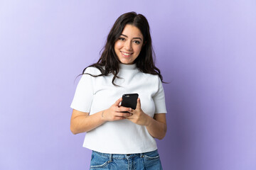 Wall Mural - young caucasian woman isolated on purple background sending a message with the mobile