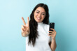 © luismolinero - Young woman using mobile phone isolated on blue background smiling and showing victory sign