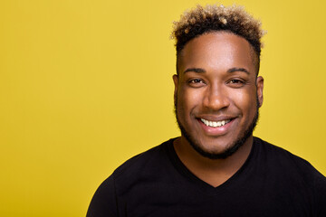 Wall Mural - Curly African-American shines with a snow-white smile after the dentist. Young stylish black man in an everyday black T-shirt smiles against a yellow background at a close-up camera. 