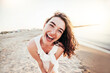 Young joyful woman in white shirt smiling at camera on the beach - Traveler girl enjoying freedom outdoors on a sunny day - Wellbeing, healthy lifestyle and happy people concept