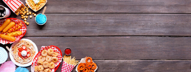 Carnival theme food corner border over a dark wood banner background. Overhead view with copy space. Summer fair concept. Corn dogs, funnel cake, cotton candy and snacks.