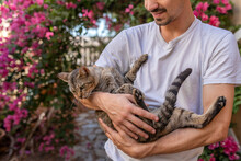 Young Man Holds In His Arms A Gray Tabby Cat With Green Eyes In The Garden