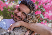 Close Up. Young Man Hugs A Gray Tabby Cat With Green Eyes In The Garden And Smiles