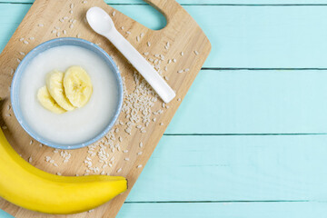 Wall Mural - Rice porridge for a baby from ground cereals in a blue bowl with a banana on a wooden board on a mint background. The first complementary feeding of the child. Place for text, recipe.