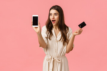 Wall Mural - Amused and surprised, good-looking caucasian woman in dress, holding credit card and showing something stunning and cool at smartphone screen, standing pink background joyful