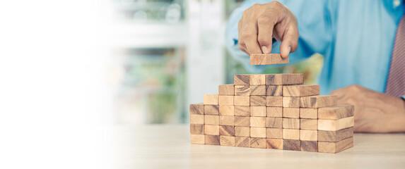 Hand placing wooden block tower stack in pyramid stair step with caution to prevent collapse or crash concepts of financial risk management and strategic planning and business challenge plan.