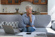 © fizkes - Confused old lady retiree sit on sofa with sad upset look distracted from financial papers debts unpaid taxes think what to do. Distressed worried senior female having no money to pay overdue bills