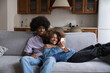 © fizkes - Cheerful dating teen Black couple using online app on smartphone at home. Happy young African girlfriend and boyfriend relaxing on couch, laughing, smiling, shopping on Internet on mobile phone
