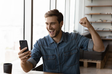 Happy millennial man in casual excited with good news, using smartphone, looking at screen, making winner yes gesture, shouting for joy. Guy reading text message, celebrating win, high result