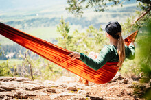 Woman With Cap Resting In Comfortable Hammock In Mountains. Relaxing On Orange Hammock Between Two Trees Pine Enjoying The Nature View