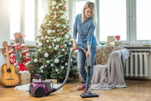 Young Woman Cleaning With Vacuum Cleaner, Vacuuming Under Christmas Tree Needles With New Years Ornaments On Hardwood Wooden Floor.