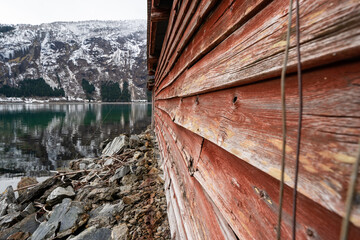 Facade of an old wooden building of Scandinavian type with red wooden boards, where there is a stone wall at the bottom behind which you can see the water