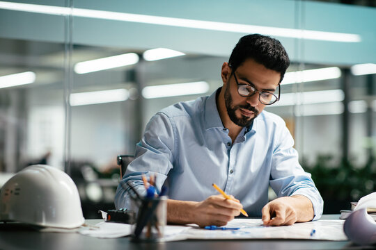 serious attractive millennial arabic man engineer in glasses with beard works with drawings at workp