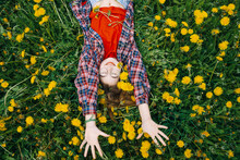 A Young Girl In A Plaid Shirt Is Lying In A Field Surrounded By Yellow Dandelions, Smiling And Pulling Her Hands Up