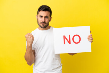 Wall Mural - Young handsome caucasian man isolated on yellow background holding a placard with text NO and angry