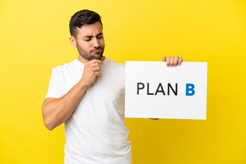 Wall Mural - Young handsome caucasian man isolated on yellow background holding a placard with the message PLAN B and thinking