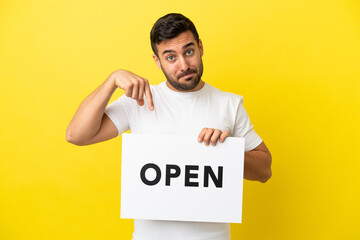 Wall Mural - Young handsome caucasian man isolated on yellow background holding a placard with text OPEN and  pointing it