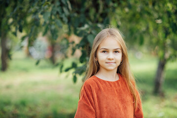 Close up portrait of beautiful little girl with blonde long hair and big blue eyes looking at camera with relaxed expression. Nature background.