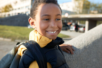 Wall Mural - Adorable male child smiling at camera over blurred background of city landscape , going to school in sunny morning, carrying black satchel on shoulders, dressed in yellow hoodie and puffer vest