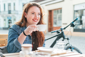 Wall Mural - Portrait of sad red curled long hair caucasian teen girl sitting on a cozy cafe outdoor terrace on the street and drinking coffee. Young woman taking a break in her city bicycle tour.