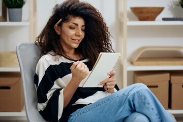 Concentrated pensive pretty beautiful curly tanned Latin woman with pen in hair in striped shirt hold notebook look down read work letter essay in home interior. Copy space Banner. Writer concept