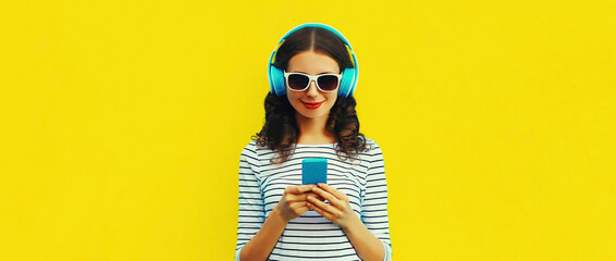 Wall Mural - Portrait of smiling young woman in headphones listening to music with smartphone on yellow background, blank copy space for advertising text