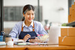 © David - Young Asian entrepreneur SME woman checking product on stock take note with a laptop. Small business owner working at home. Check address on the parcel SME online start up freelance concept.