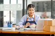 © David - Young Asian entrepreneur SME woman checking product on stock take note with a laptop. Small business owner working at home. Check address on the parcel SME online start up freelance concept.
