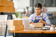 © David - Young Asian entrepreneur SME woman checking product on stock take note with a laptop. Small business owner working at home. Check address on the parcel SME online start up freelance concept.